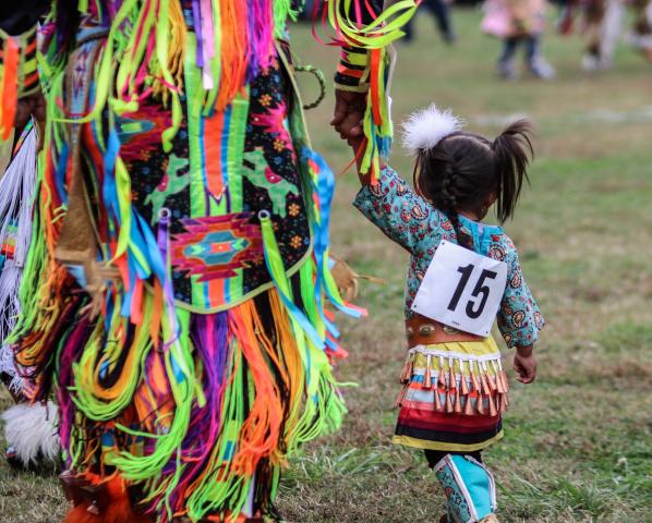 A young girl with jingle bells holds her mother's hand. Her father said she's been dancing at powwows "ever since she could walk." 